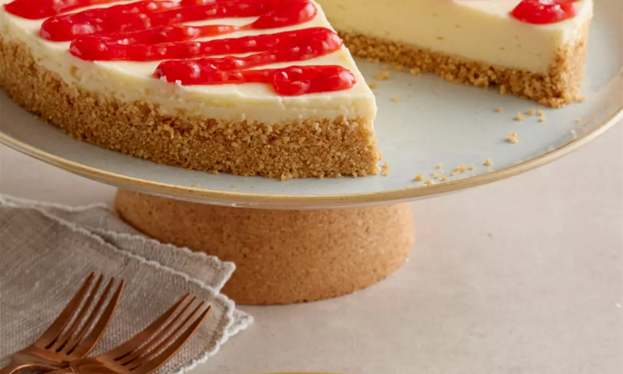 Galeta unlu cheesecake altın rengi tabanlı, kremsi beyaz dolgu üzerinde parlak kırmızı sos şeritleriyle sade bir tabağa yerleştirilmiş