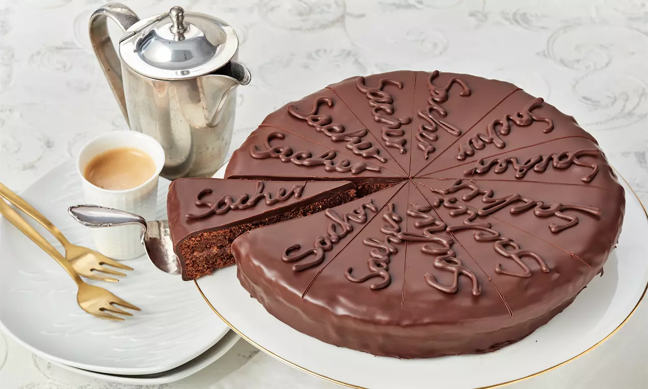 Tamamıyla çikolata kaplı çember şeklindeki Sachertorte, dilimlenmiş ve çikolata yazılarla süslenmiş, yanına kahve ve çaydanlık eklenmiş