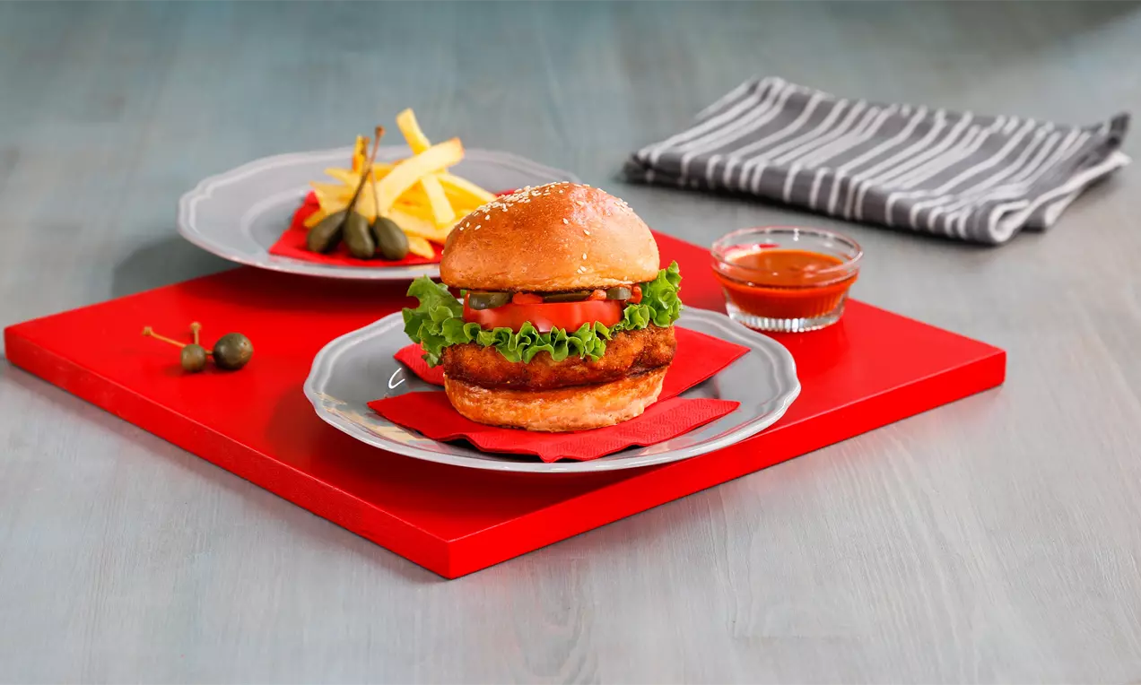 Susam kaplı altın rengi burger ekmeği içinde çıtır tavuk, yeşil marul ve dilimlenmiş domates, yanında patates kızartması ve sos ile sunulmuş
