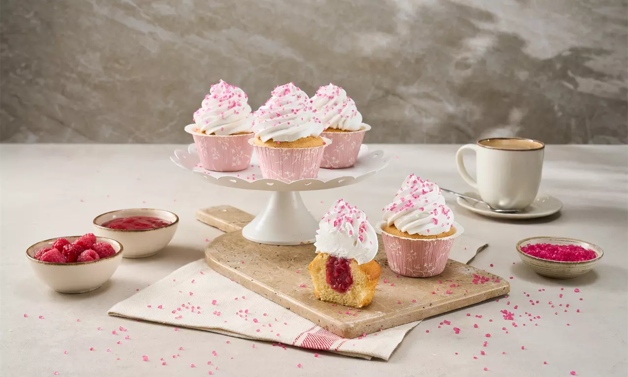 Pembe kağıt kaplarda krema ve pembe şeker kristalleri ile süslenmiş cupcake’ler, ahşap sunum tahtası ve beyaz bir pasta standında sergilenmiş