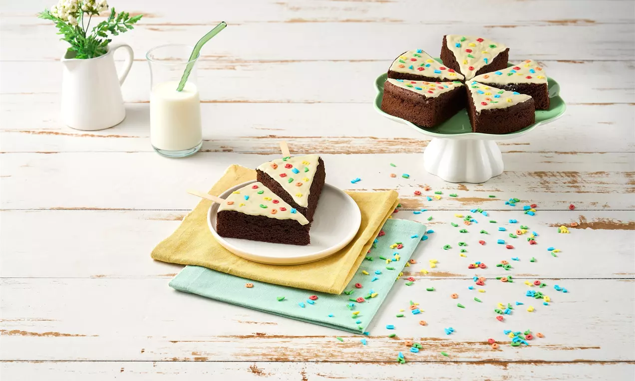 Kakao bazlı kek dilimleri, beyaz krema kaplı, renkli şekerleme serpiştirilmiş, pastel peçetelerle servis edilen beyaz tabakta.