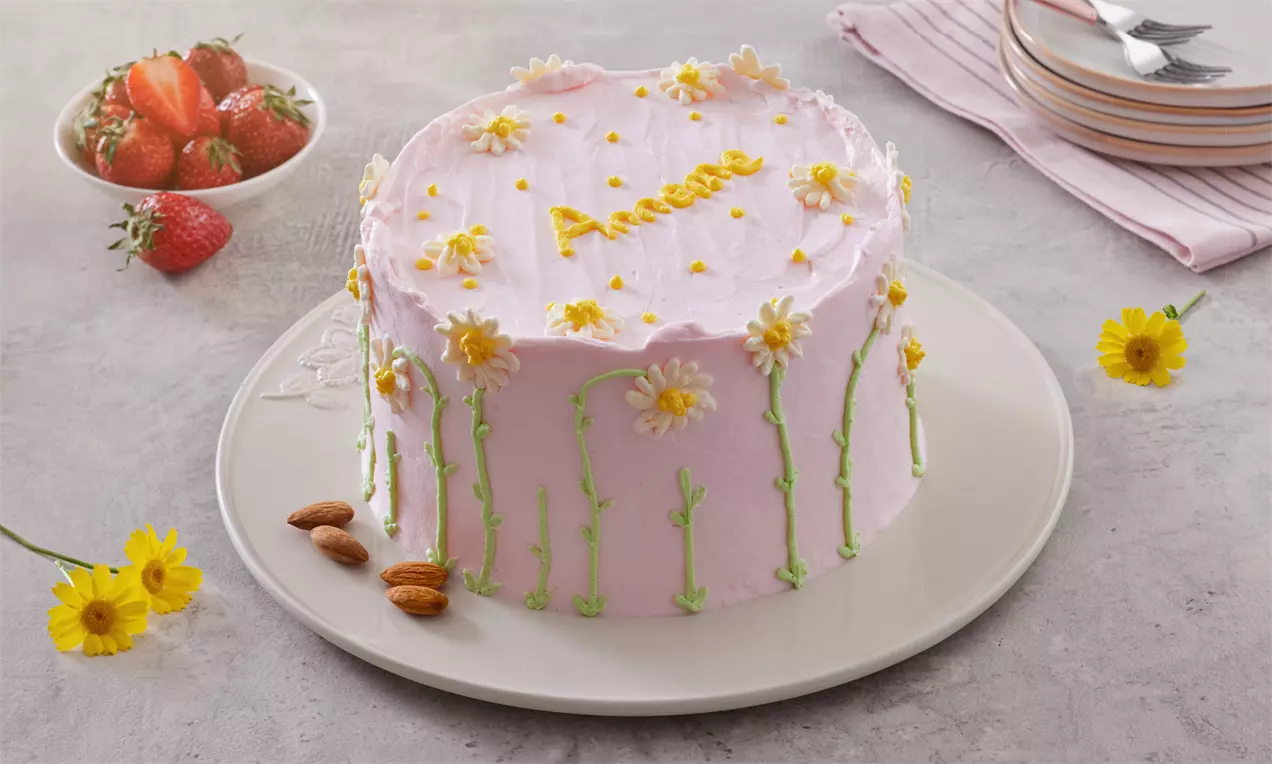 Pembe krema kaplı yuvarlak pasta, sarı papatyalar ve yeşil yaprak desenleriyle süslenmiş, beyaz tabakta badem ve çileklerle sunulmuş