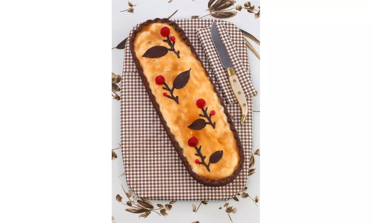 Kahverengi tart tabanı içinde kremalı dolgu, kırmızı meyve ve çikolata yapraklarıyla süslenen dikdörtgen tatlı, kareli bir kumaş üzerinde sergileniyor