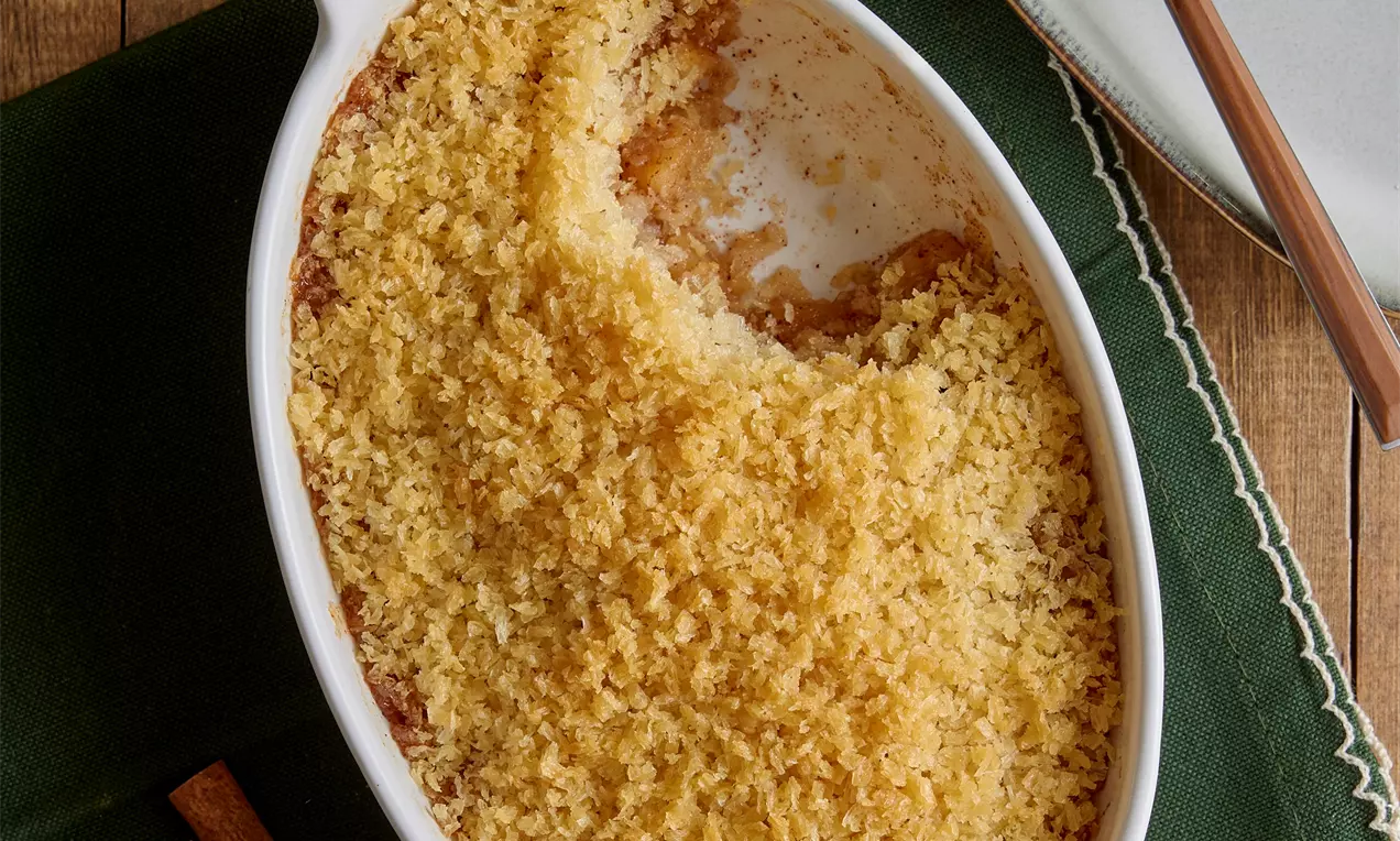 Oval bir beyaz fırın kabında altın sarısı pane ile kaplanmış elmalı tatlı, yanında yeşil kumaş ve tabakta sunulan bir porsiyon