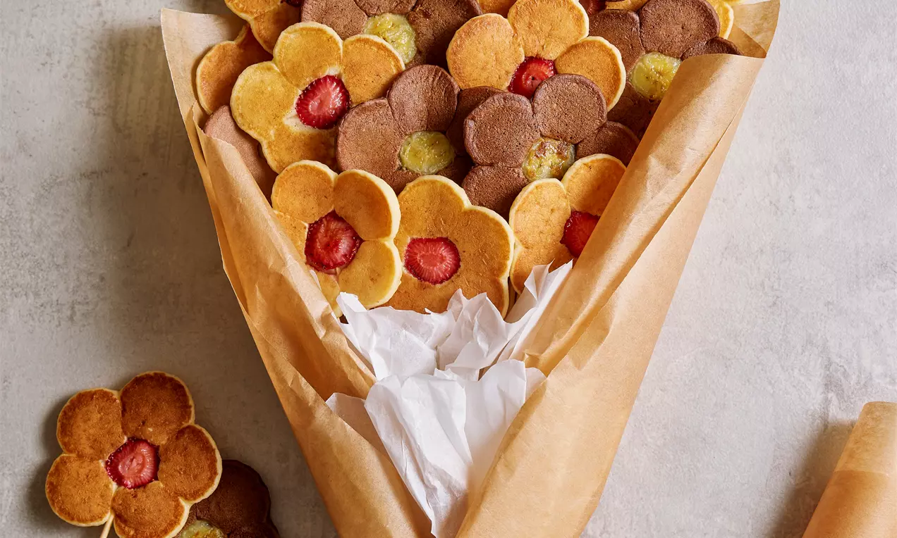 Kahverengi ve altın sarısı çiçek şekilli pancake’ler çilek ve kivi dilimleriyle süslenmiş, kraft kağıda sarılı ve beyaz kurdele ile bağlanmış