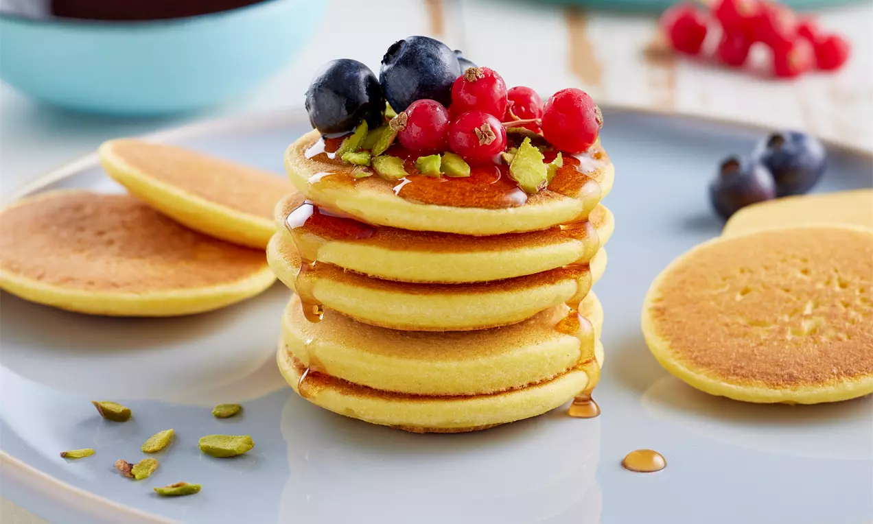 Altın sarısı mısır unlu pancake'ler üst üste dizilmiş, üzeri meyveler ve Antep fıstığıyla süslenmiş, yanlarda soslu ve sade pancake'ler bulunuyor