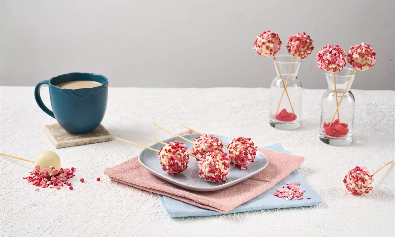 Beyaz çikolata kaplı lolipop kekler pembe ve kırmızı parçacıklarla süslenmiş, pastel tonlarda bezemelerle servis edilmiş