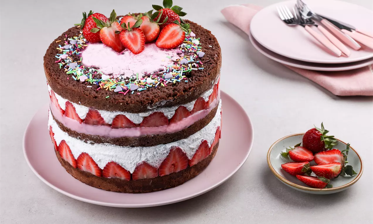 Kakao bazlı katmanlar arasında beyaz krema, dilimlenmiş çilekler ve pembe dolgu, üstte çilek ve renkli serpiştirmelerle süslü