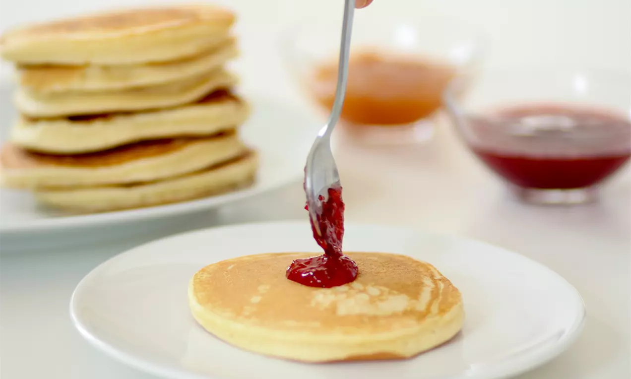 Altın sarısı, yumuşak dokulu pancake bir beyaz tabakta, üzerine kaşıkla dökülen parlak kırmızı reçelle görsel bir uyum oluşturuyor