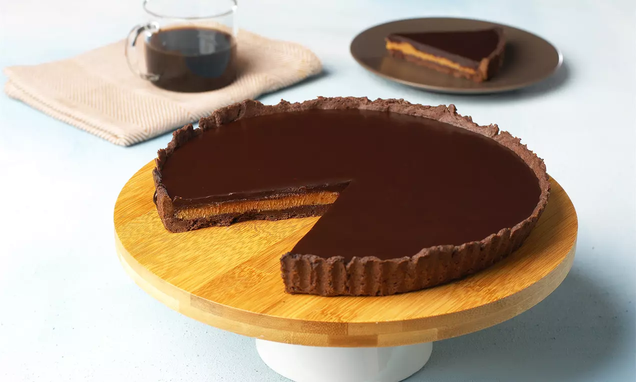 Koyu parlak çikolata kaplı, dalgalı kenarlı yer fıstıklı tart ahşap bir stant üzerinde, yanında kahve ve dilim tabağıyla sunulmuş
