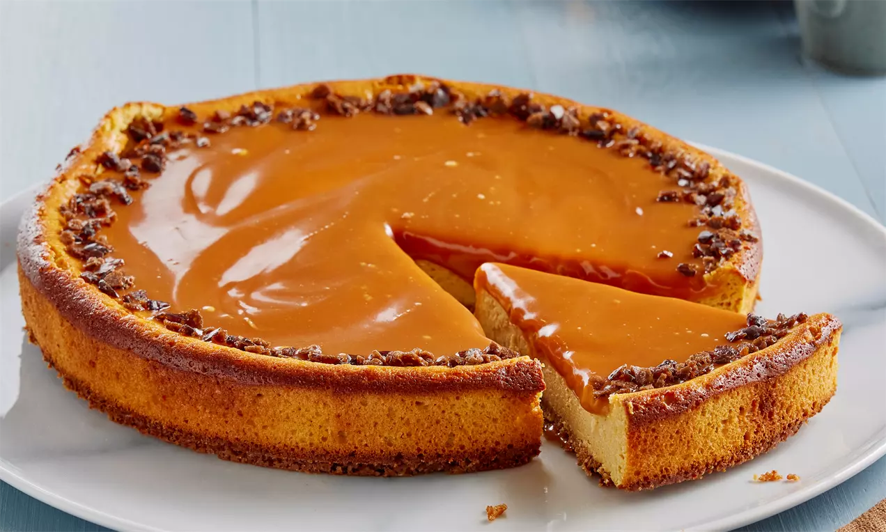 Yuvarlak beyaz tabakta sunulan, karamel dolgusuyla kaplanmış çıtır bisküvi kenarlı tereyağlı cheesecake, dilimlenmiş halde