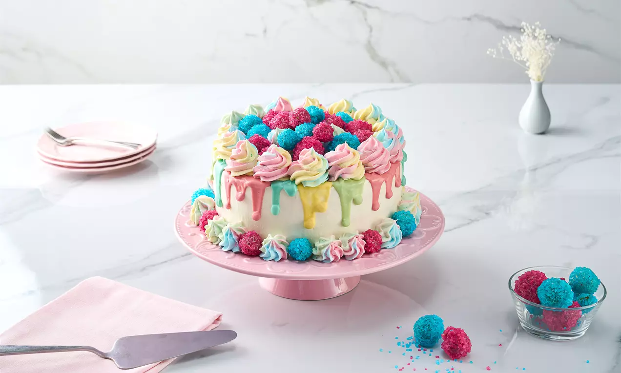 Pastel tonlarda krem parlak damlalarla süslenmiş, renkli kristalimsi toplarla çevrili ve pembe bir tabakta sunulan katmanlı pasta